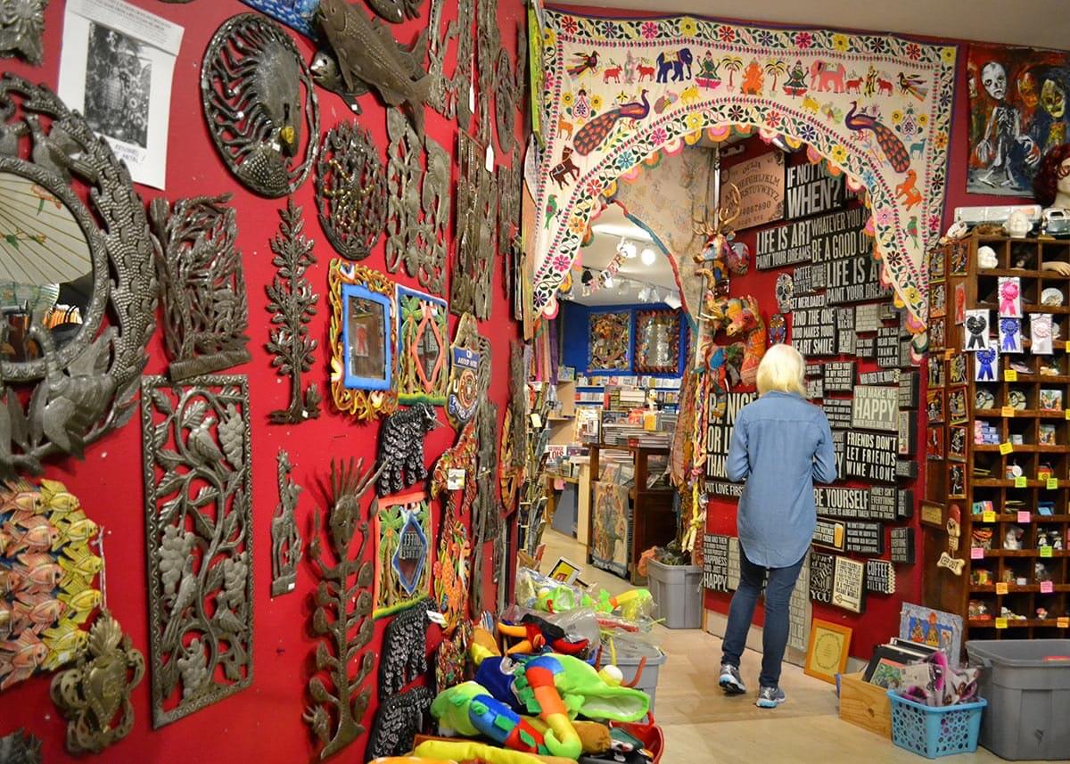 A blonde woman walking through a hallway full of eclectic museum pieces hanging
                on the walls, such as floral bronze carvings, an embroidered curtain with colorful birds and a Ouija
                board.