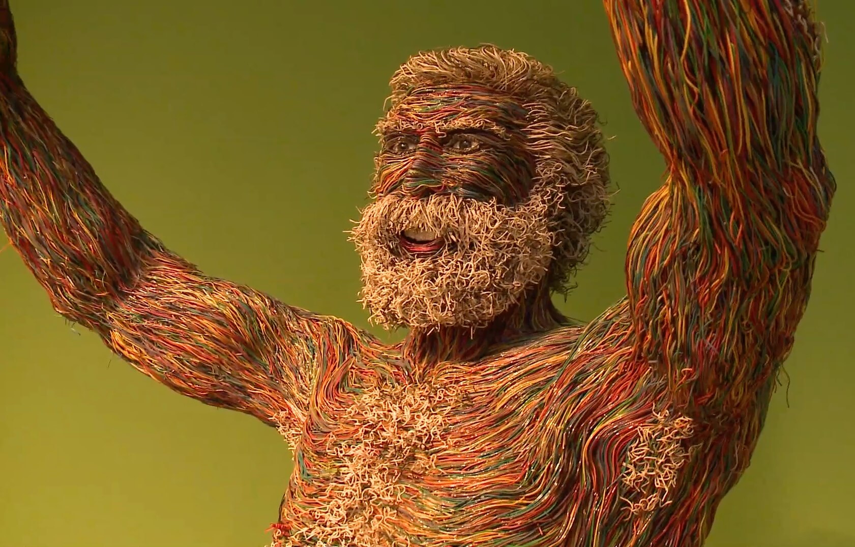 A naturalistic sculpture of a bearded old man with his arms raised and smiling,
                composed entirely out of red, green and yellow wires, except for his white-wired beard and hair.